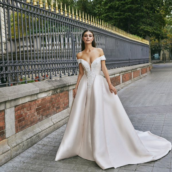 Thumbnail of a brunette model stood by black railing in Ronald Joyce 69713, a light gold fit and flare off-the-shoulder Mikado wedding dress with a plunging neckline and A-line overskirt